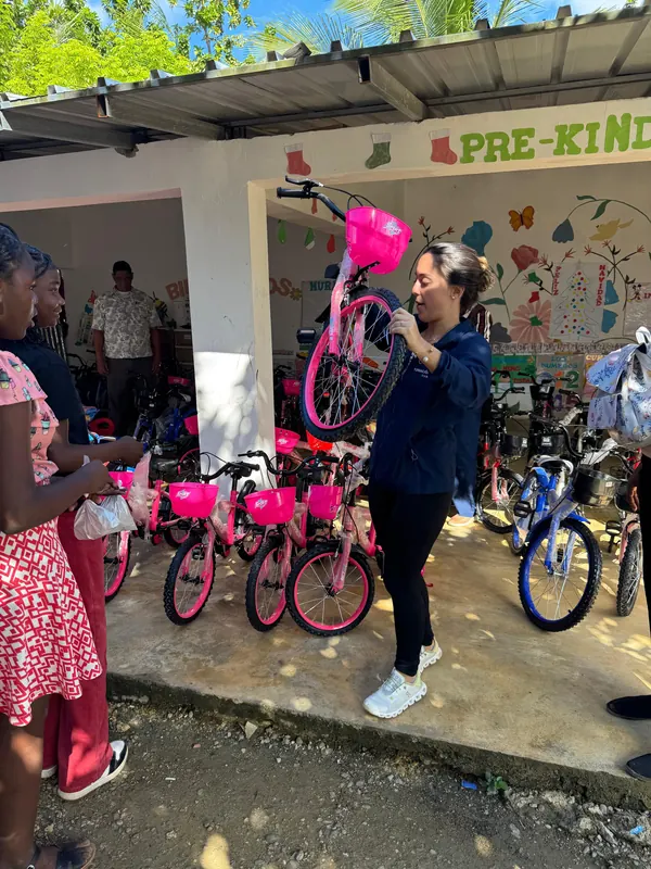 Una voluntaria carga una bicicleta rosada nueva a la entrada de un aula de pre-kínder, frente a las demás bicicletas.