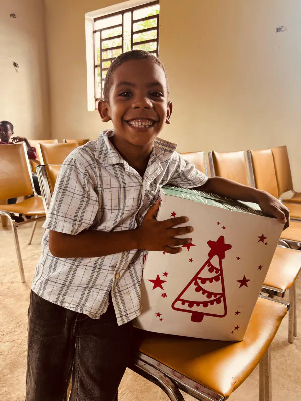 Un niño sonríe abrazando una caja de regalo blanca decorada con un árbol de Navidad rojo.