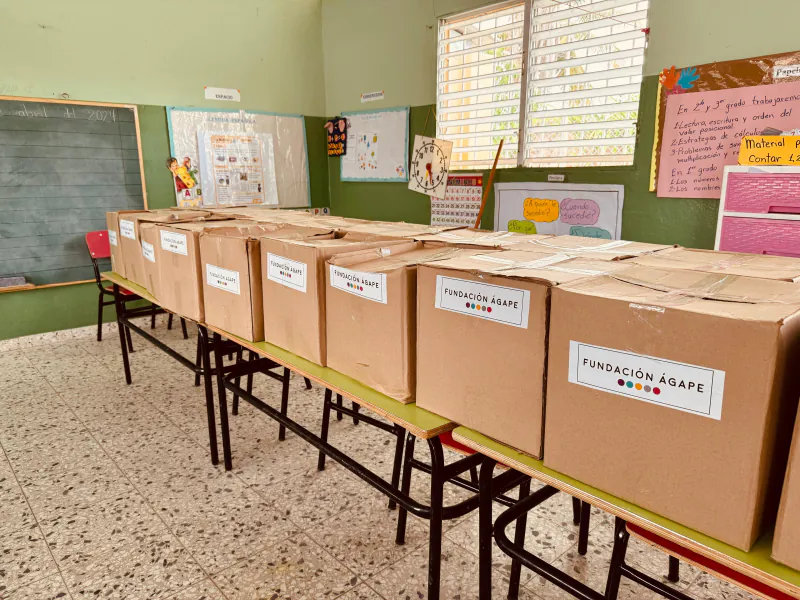 Fila de cajas de cartón rotuladas «Fundación Ágape» sobre los pupitres de un aula, listas para entregar.