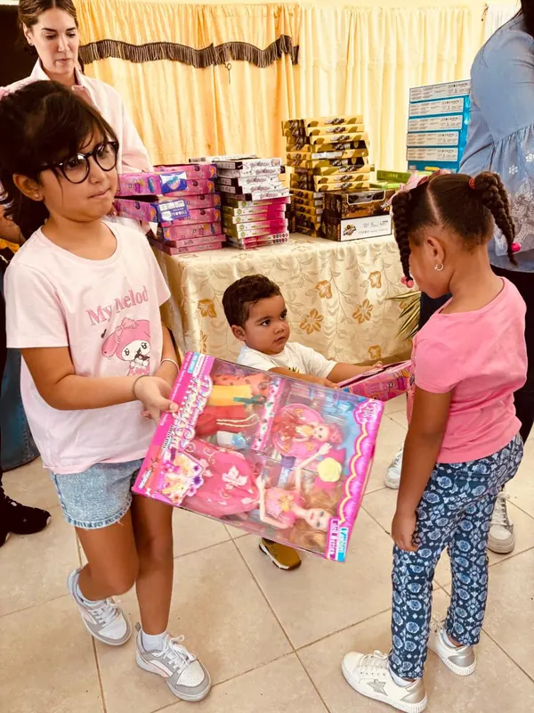 Dos niñas reciben juguetes de manos de una voluntaria, junto a una mesa cubierta de cajas de regalos.