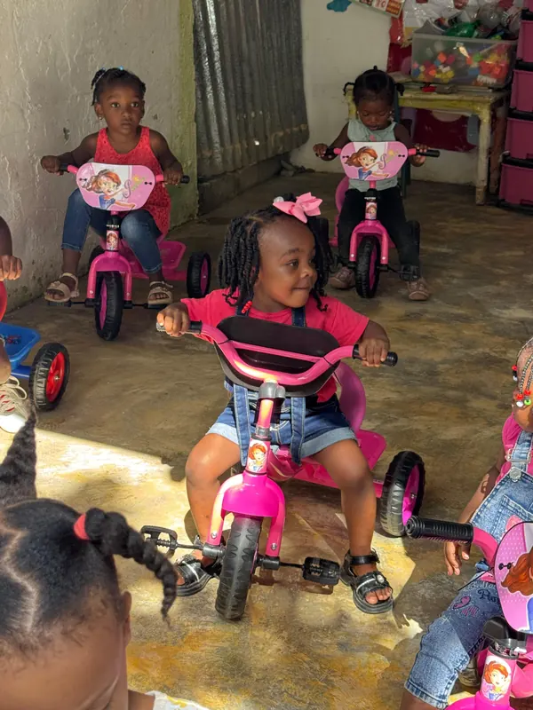 Dos niñas pedalean triciclos rosados recién entregados dentro de un aula de nivel inicial.
