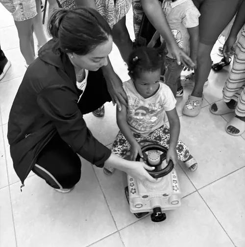 Fotografía en blanco y negro: la fundadora se agacha para ayudar a una niña pequeña a montarse en un carrito de juguete.