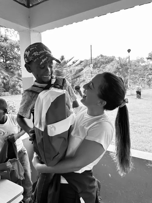 Fotografía en blanco y negro: la fundadora ríe mientras carga en brazos a un niño que lleva puesta su mochila nueva.