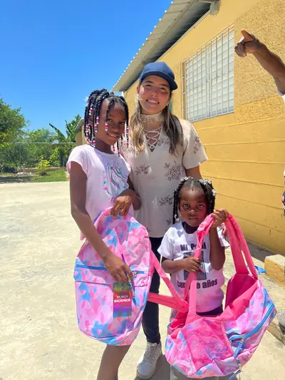 Una voluntaria acompaña a dos niñas que muestran sus mochilas nuevas frente a una escuela.
