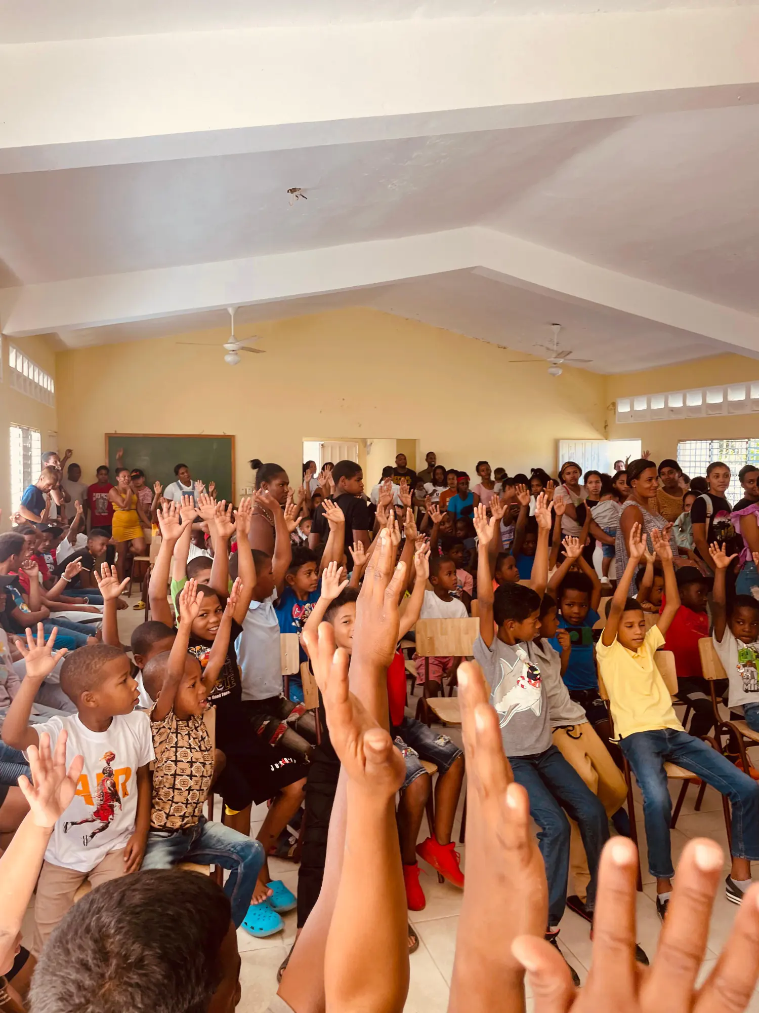 Un salón lleno de niños y niñas sentados que levantan las manos, con voluntarias al frente, durante una jornada de la fundación.