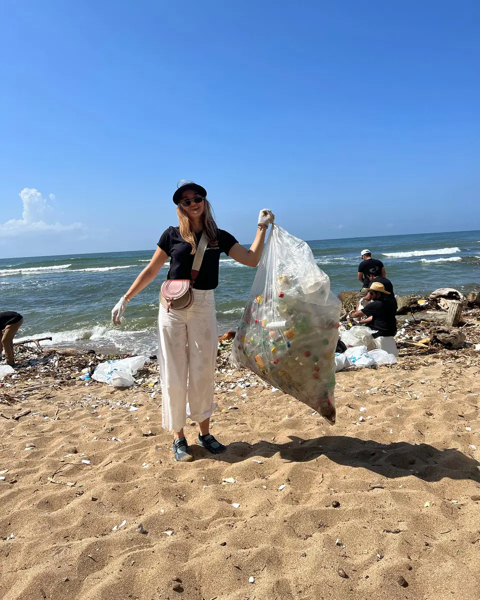 Una voluntaria sonríe frente al mar sosteniendo en alto una funda transparente llena de botellas y plásticos recogidos de la arena, mientras el resto del equipo sigue trabajando detrás.