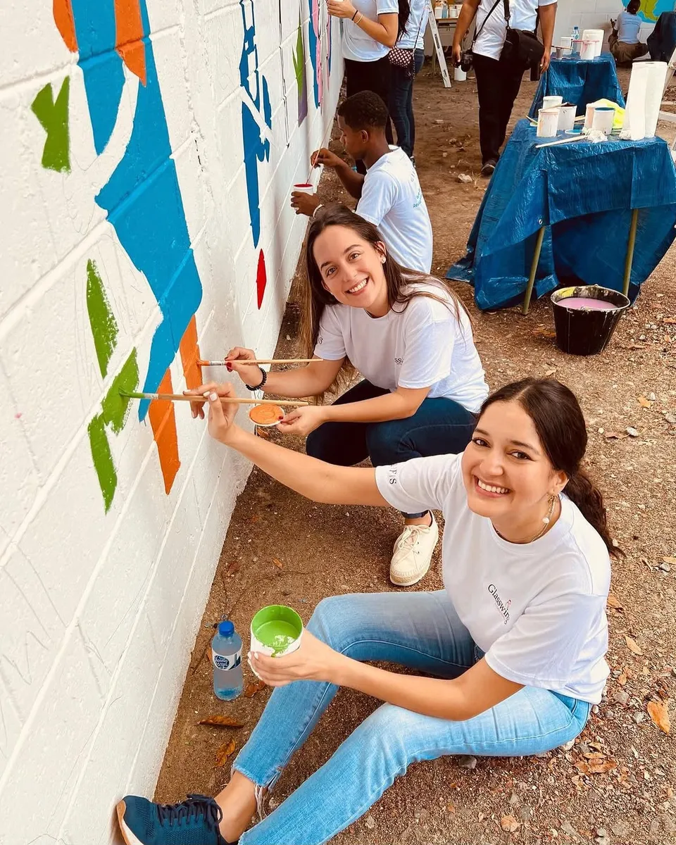 Dos voluntarias sonríen a la cámara mientras pintan con pinceles un mural de colores en el muro de un centro comunitario, con más voluntarios trabajando detrás.