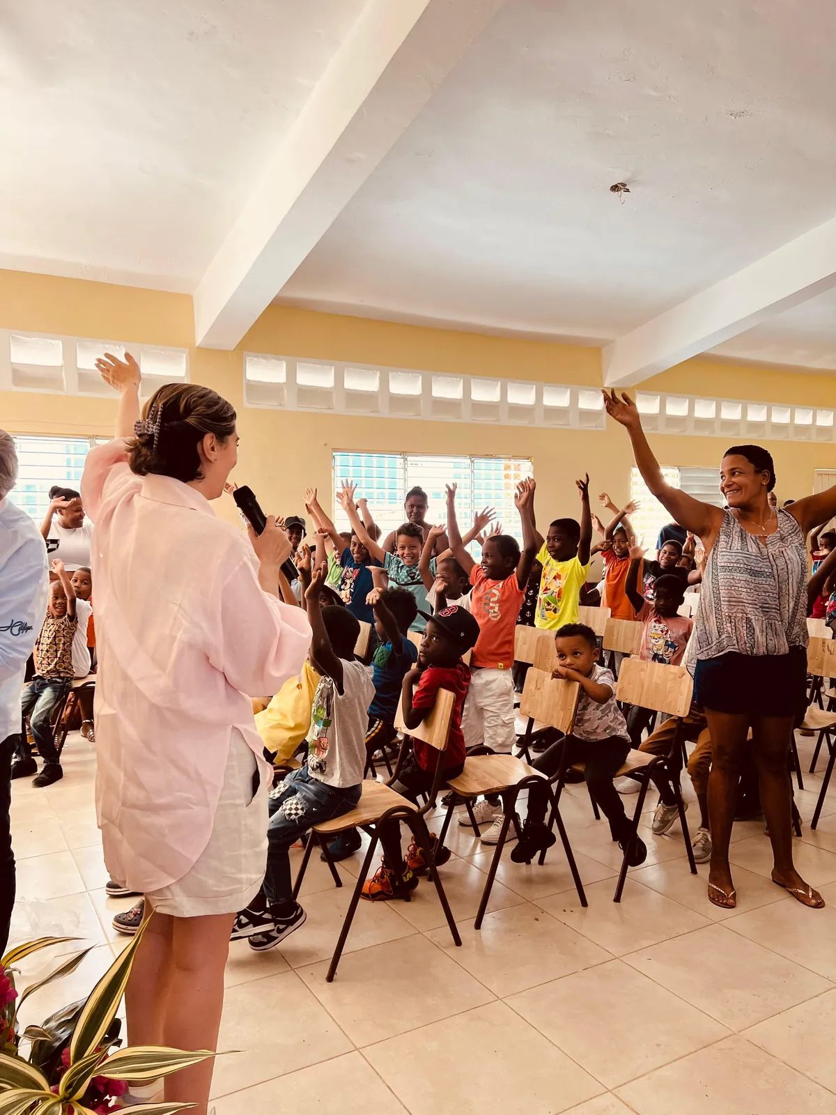 Una voluntaria con micrófono anima una actividad en un salón lleno de niños y niñas que levantan la mano para participar.
