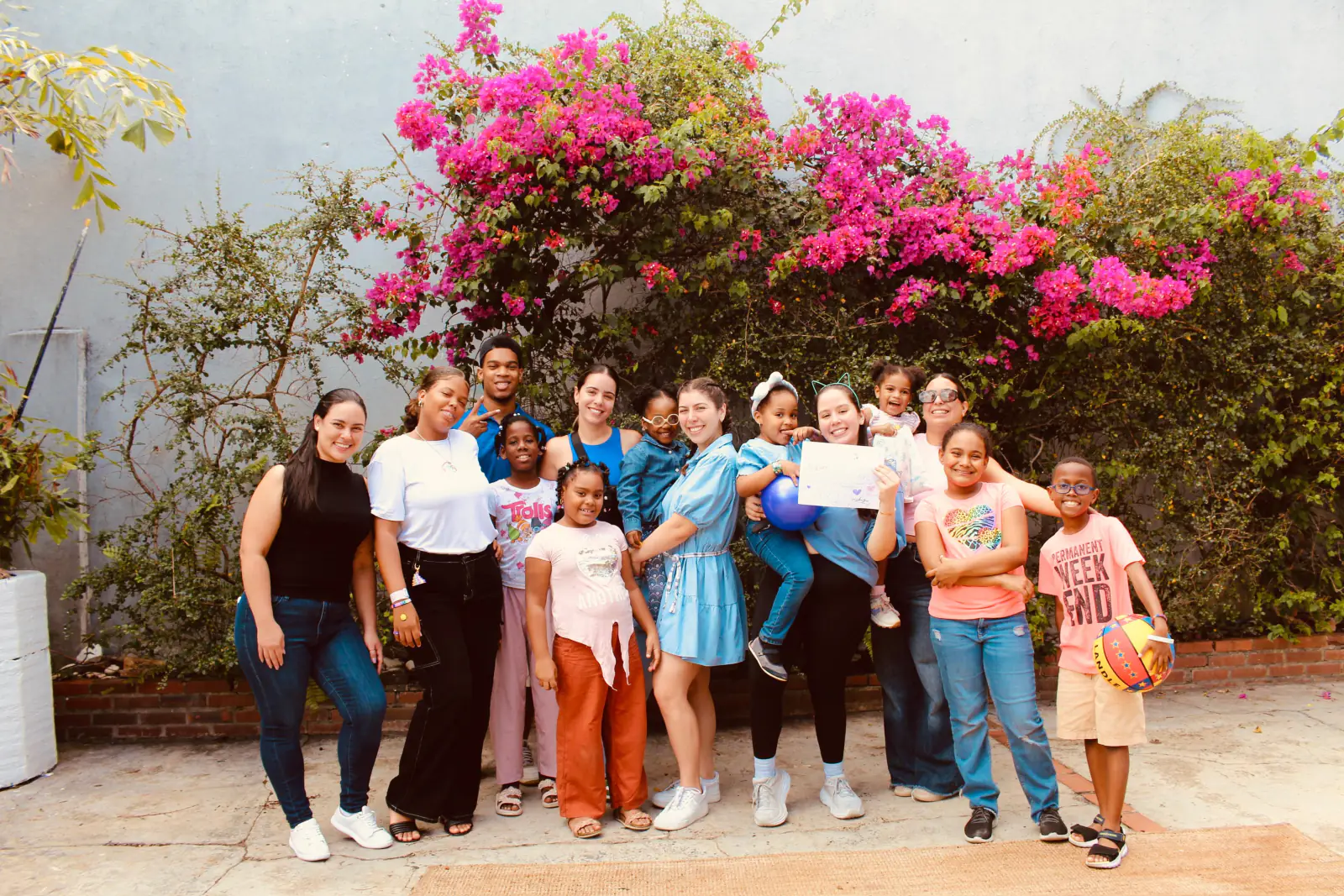 Un grupo de voluntarias y voluntarios posa junto a un grupo de niños y niñas frente a una buganvilia en flor, al cierre de una jornada.